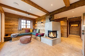A log cabin interior with a stone fireplace, a seating area with cushions, and a large window. - Alm Kone Chalet, 130 m² for 4 people, wood-burning stove, pocket-spring mattresses (Samerberg)