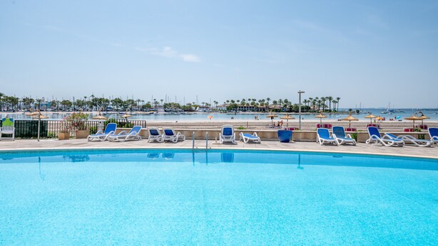 A swimming pool with lounge chairs, a beach in the background, and a clear sky. - Vacation Apartment “Carabela Dali 113” Close to the Beach with Sea View and Pool (Alcudia)