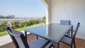 A patio with a glass table and chairs, overlooking a beach with umbrellas and palm trees. - Vacation Apartment “Carabela Dali 113” Close to the Beach with Sea View and Pool (Alcudia)