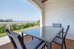 A patio with a glass table and chairs, overlooking a beach with umbrellas and palm trees.