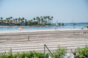 A beach with a fence, palm trees, and boats in the water.