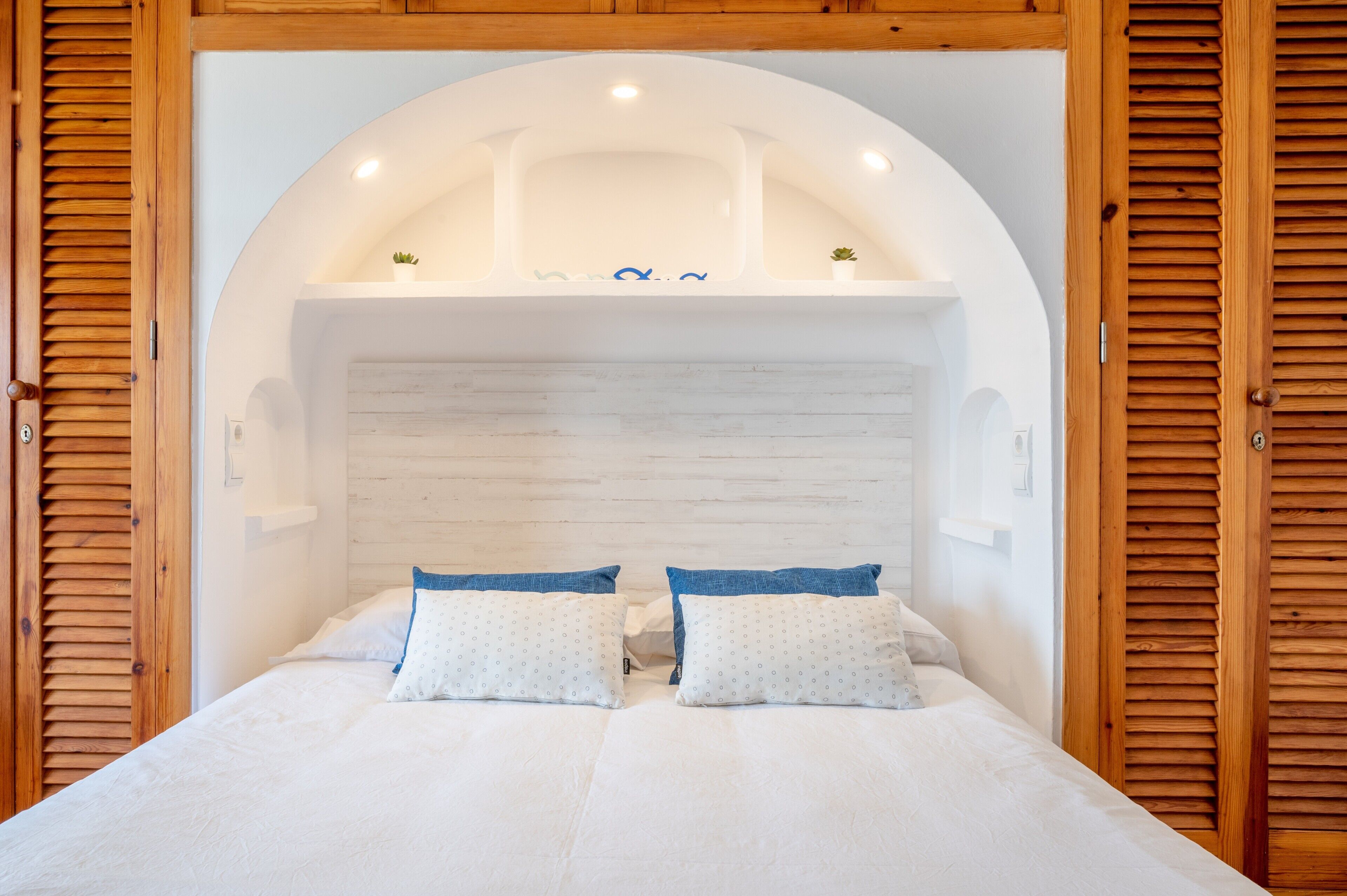 Un dormitorio con cama blanca, dos almohadas, cabecera de madera y una pequeña estantería decorativa.