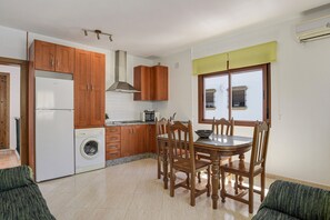 Une cuisine avec des meubles de rangement en bois, un lave-linge, une table à manger avec des chaises et une fenêtre avec des stores.