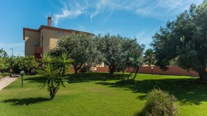 A building with a flat roof, a chimney, and a garden with grass, trees, and a small bush.