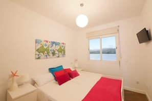 A bedroom with a bed, a window with a view, a painting, and a TV on the wall.