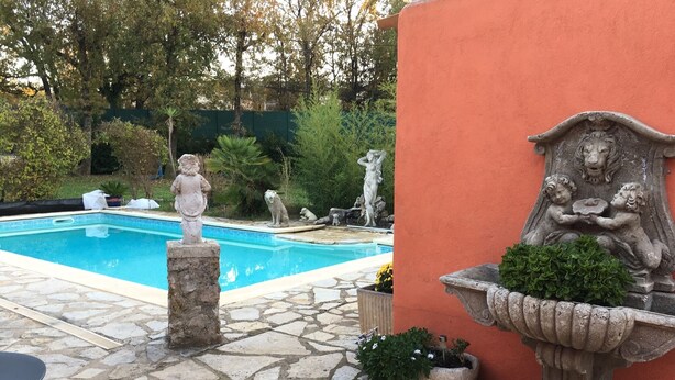 Una piscina con baldosas de piedra, rodeada de estatuas y una jardinera de piedra con follaje.