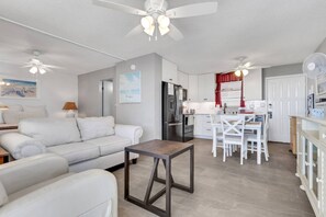 Een moderne woonkamer met een bank, een salontafel en een eethoek met een tafel en stoelen. Er is een keuken met een koelkast en een raam met rode gordijnen.