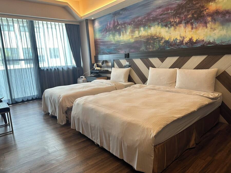 A hotel room with two beds, a large window with curtains, a painting on the wall, and a wooden floor.