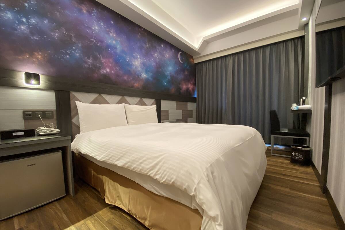 Ein modernes Hotelzimmer mit einem großen Bett, einem Schreibtisch, einem Nachttisch und einer Wandmalerei zum Thema Galaxie.