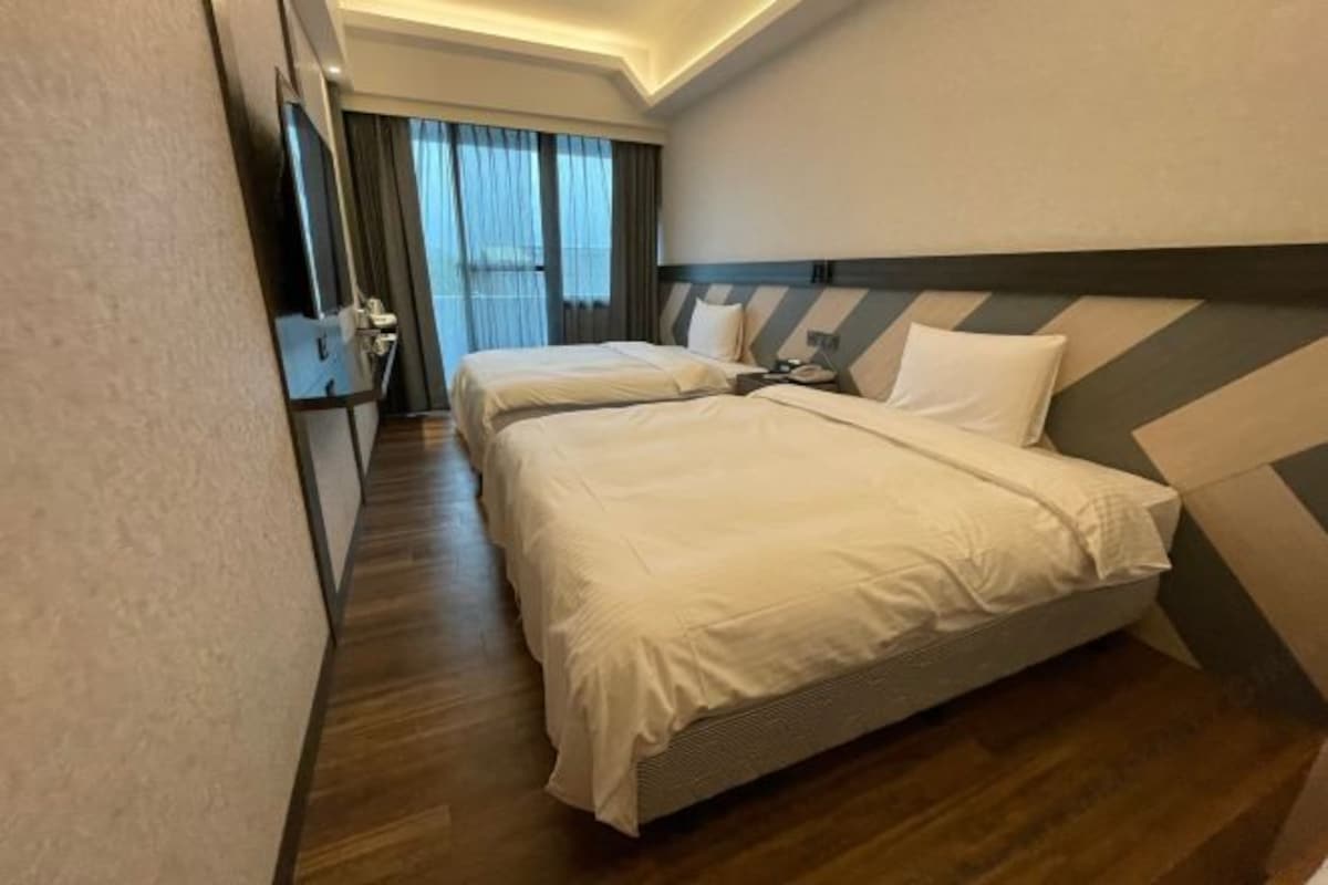 Habitación de hotel con dos camas, piso de madera, un ventanal grande con cortinas y un cabecero moderno.