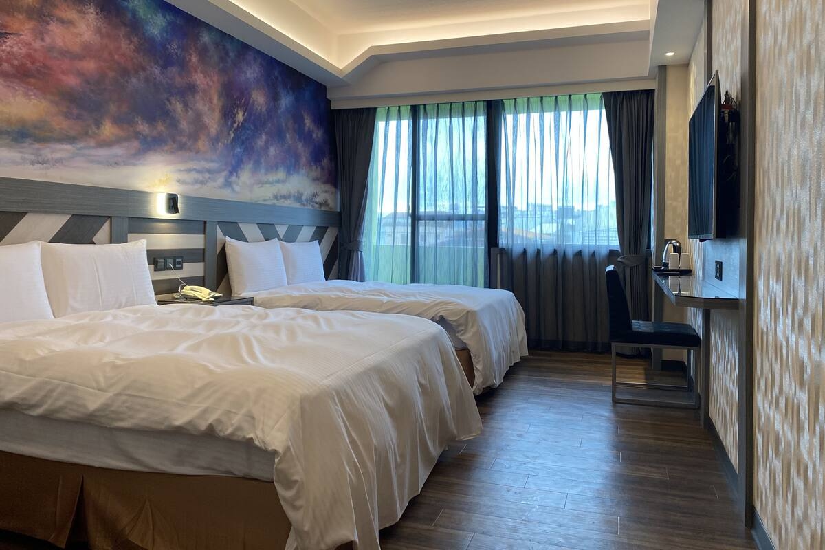 Ein Hotelzimmer mit zwei Betten, einem Schreibtisch, einem Stuhl, einem Fernseher und einem großen Fenster mit Vorhängen.