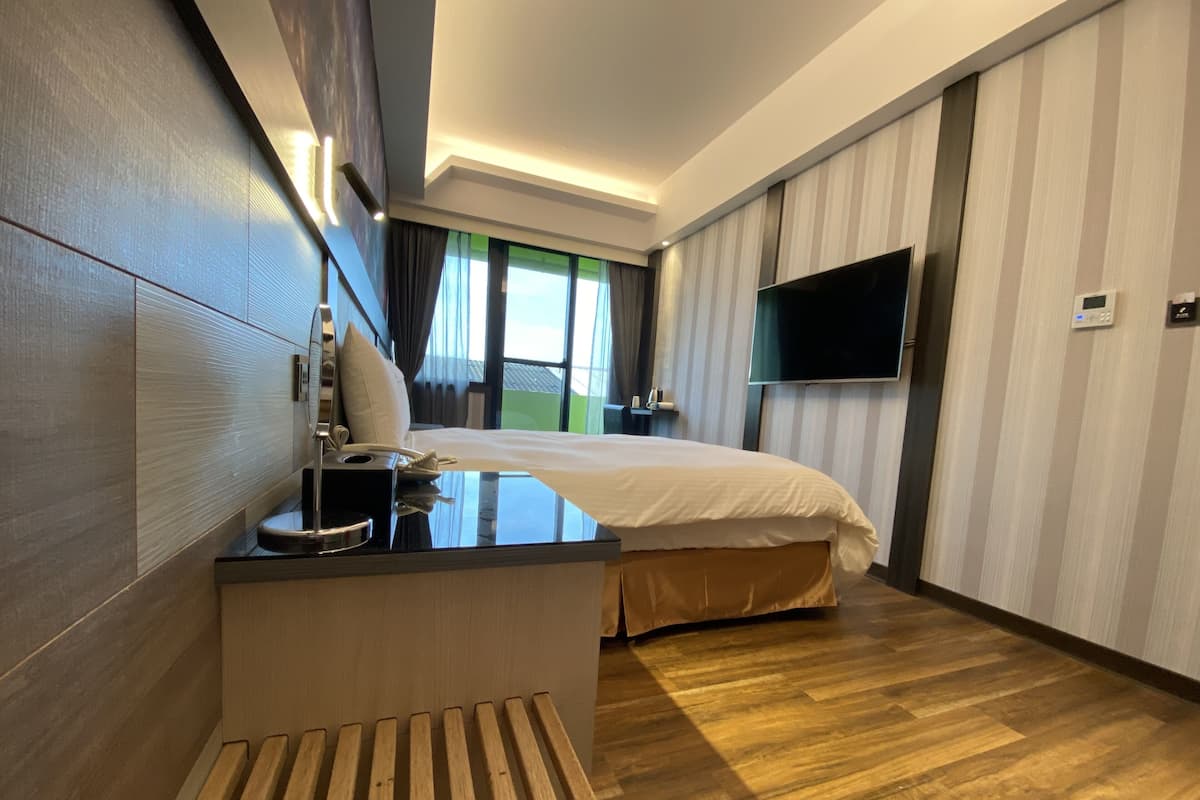 Ein modernes Hotelzimmer mit einem Holztisch, einem Bett, einem Flachbildfernseher und einem großen Fenster mit Vorhängen.