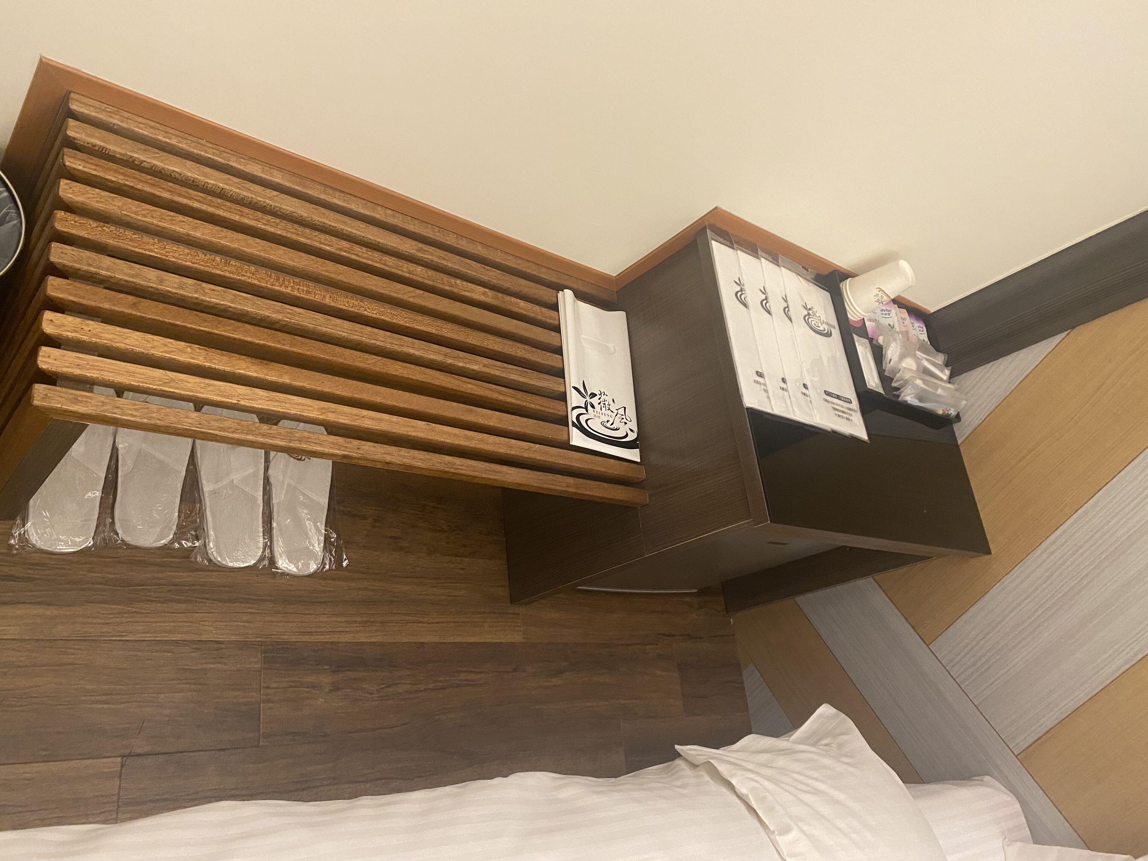 A hotel room with a wooden headboard, a bedside table with a white cloth, and a small shelf with toiletries.