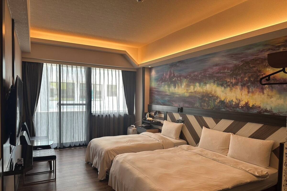 Ein Hotelzimmer mit zwei Betten, einem Schreibtisch, einem Stuhl, einem Bild an der Wand und einem Fenster mit Vorhängen.