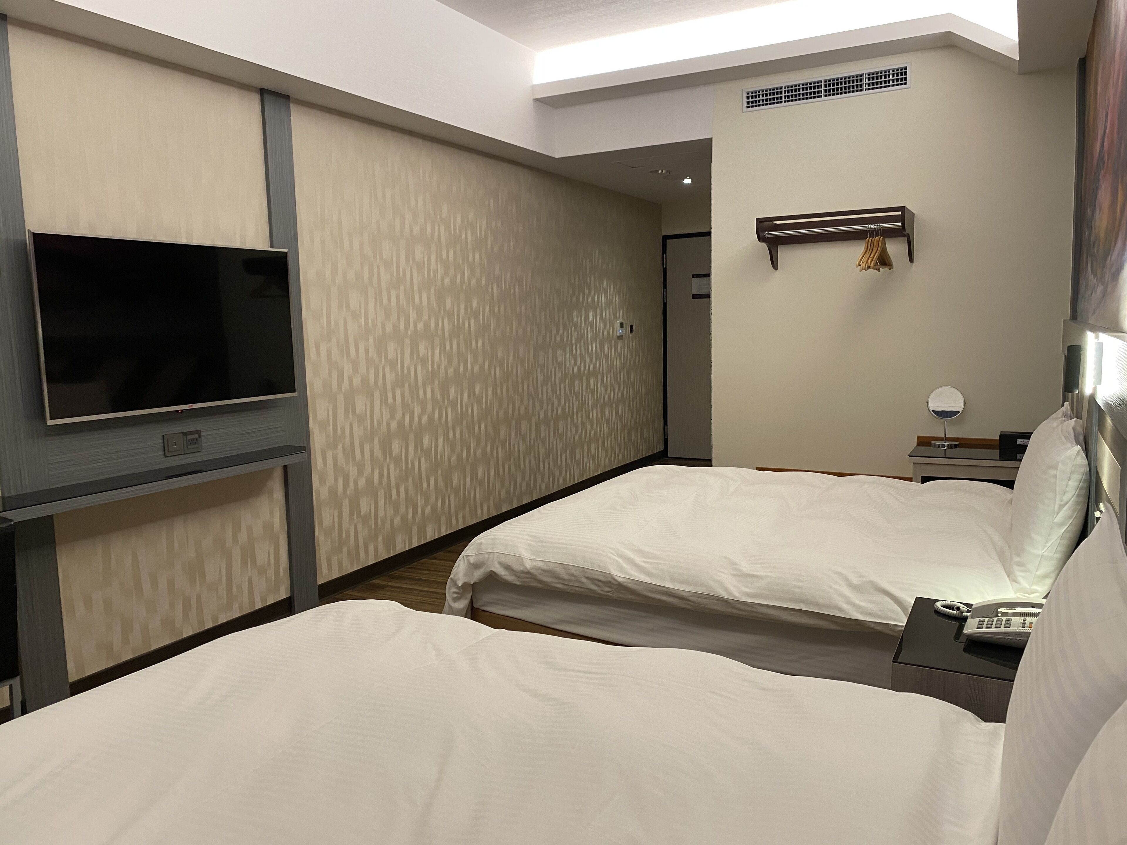 Una habitación de hotel con dos camas, un televisor de pantalla plana, una mesita de noche con lámpara y un espejo pequeño.
