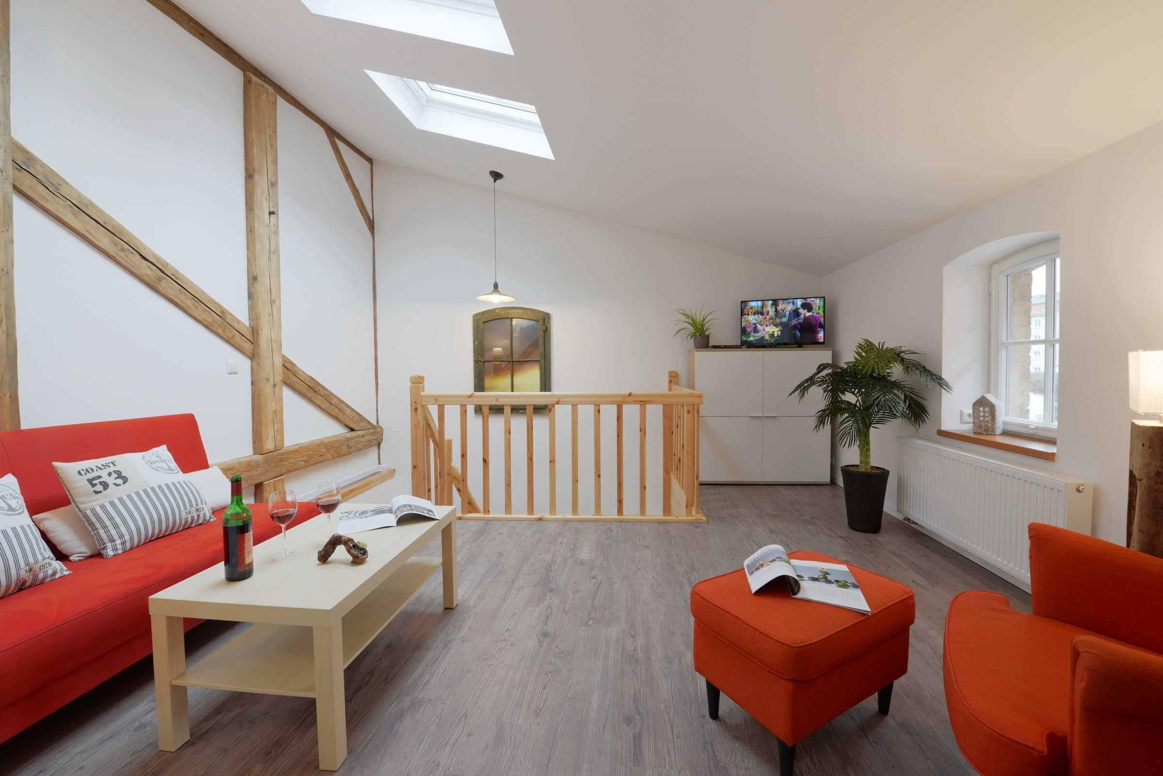 A modern living room with a red sofa, a wooden coffee table, and a small orange ottoman. There is a wooden railing and a potted plant in the background.