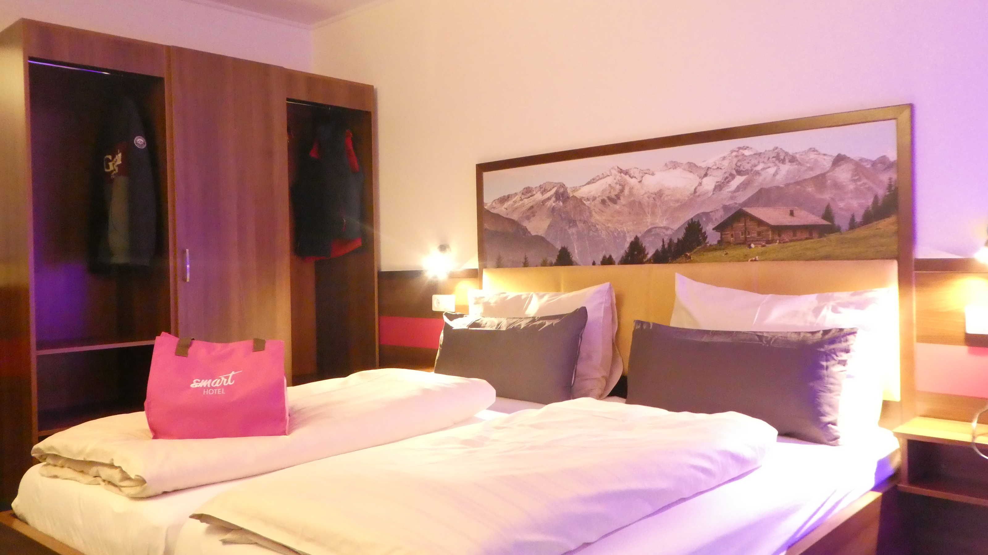 Una camera d'albergo con un letto, un armadio, un quadro raffigurante montagne e una borsa rosa sul letto.