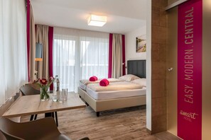 A modern hotel room with a bed, a dining table, and a view of the city.