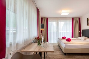 Een hotelkamer met een bed, een tafel met een vaas en glazen, en uitzicht door dunne gordijnen.