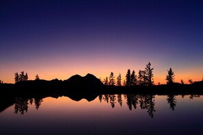 La silhouette d’une montagne se reflète dans une étendue d’eau calme, avec des arbres au premier plan.
