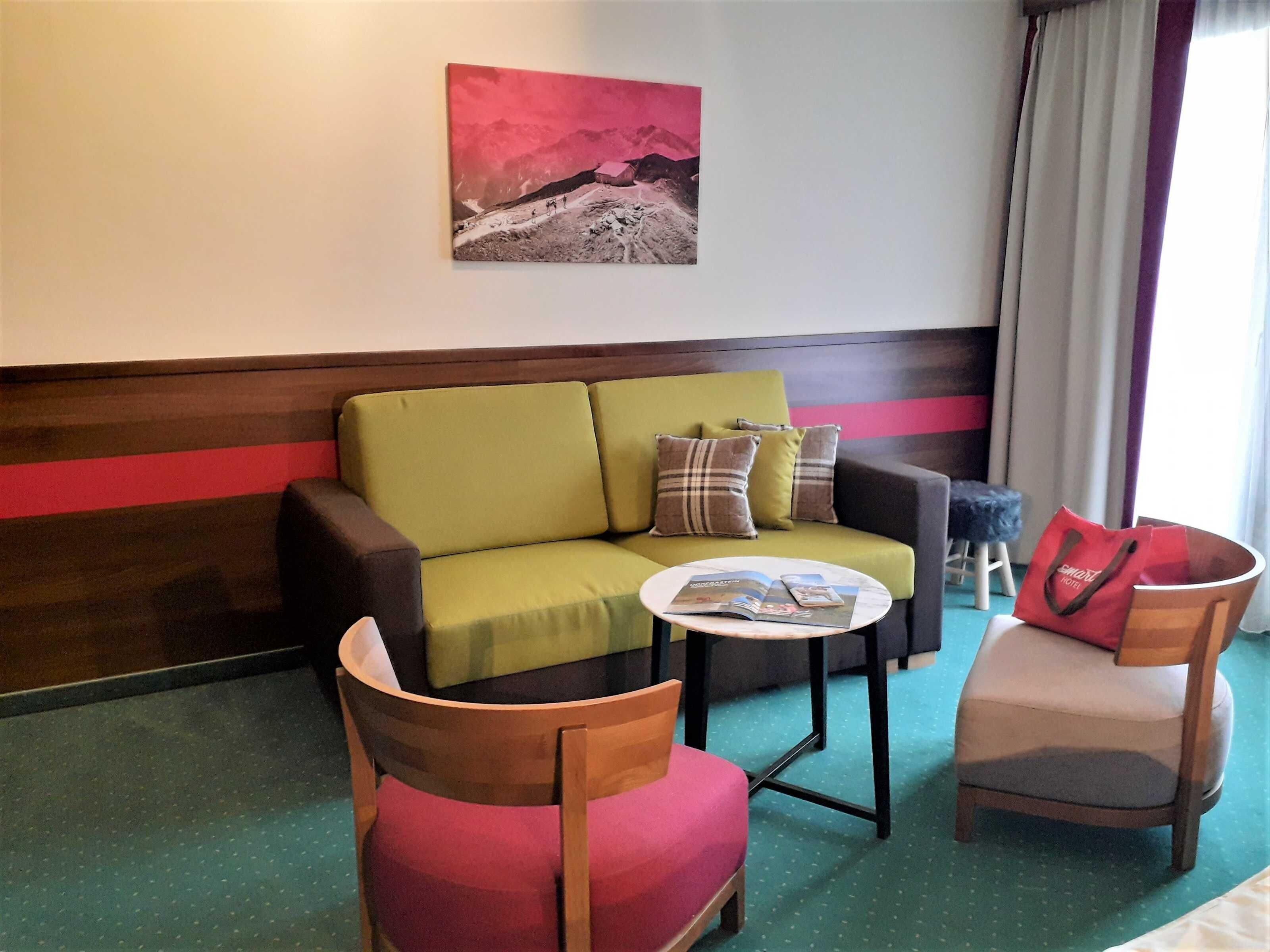 Une pièce avec un canapé vert, deux fauteuils, une petite table avec des magazines, un sac rouge et un tableau accroché au mur.