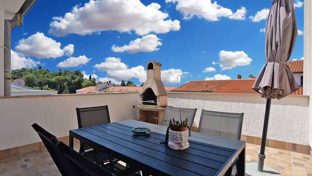 An outdoor patio with a table, chairs, a grill, and a view of the sky and buildings.