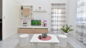 Ein modernes, minimalistisches Wohnzimmer mit weißer Farbgebung, das ein zentrales Tablett mit bunten Gegenständen, zwei weiße Stühle, einen Couchtisch, ein Bücherregal mit Dekoration und ein großes Fenster mit gestreifteren Vorhängen zeigt.