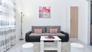 Ein modernes Wohnzimmer mit einem grauen Sofa, zwei Kissen, einem weißen Couchtisch und einem kleinen weißen Beistelltisch. An der Wand hängt ein Bild von rosafarbenen Blumen, und es steht eine hohe Holzkommode sowie ein gestreifter Vorhang im Raum.