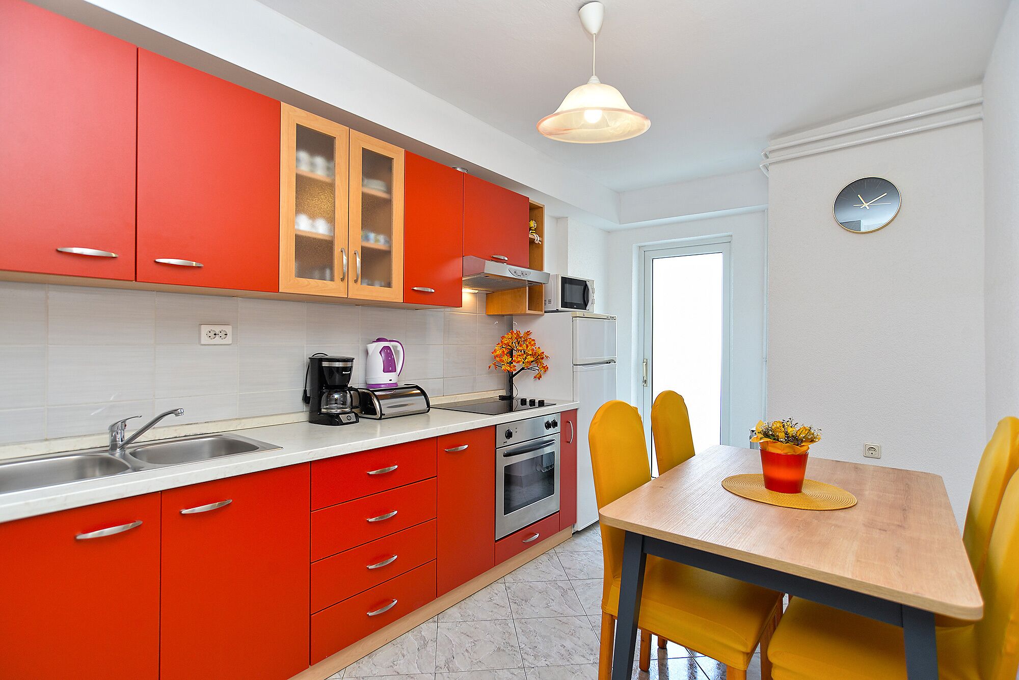 A compact kitchen with red cabinets, a white countertop, a sink, a coffee maker, a microwave, an oven, and a dining area with a wooden table and yellow chairs.
