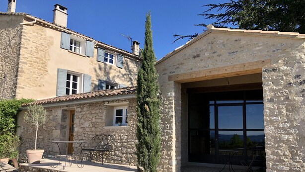 A stone building with a large garage door, a small porch with a bench, and a potted plant. - La Verrière, Très Jolie Chambre Indépendante Avec Courette Abritée (Bonnieux)