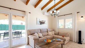 Un salón con un sofá color beige, una mesa de centro de madera y una araña. Se ve una ventana con cortinas, un cuadro en la pared y una puerta que da a un espacio exterior.