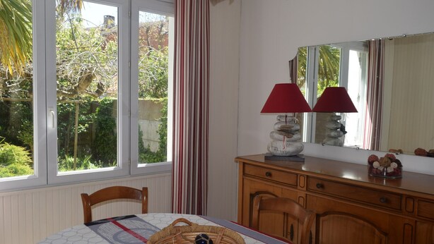A dining area with a table set for two, a wooden cabinet, and a window with a view of greenery. - Cozy Home Near Sea and Arago Market, 2-Star Comfort (Les Sables-d'Olonne)