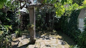 A stone-paved garden with a tree trunk, surrounded by dense foliage and a wooden fence. - Cozy Home Near Sea and Arago Market, 2-Star Comfort (Les Sables-d'Olonne)