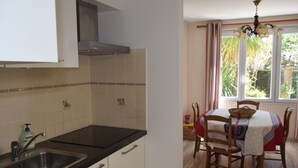 A kitchen with a stainless steel sink, white cabinets, and a black countertop. There is a dining area with a table covered by a tablecloth, wooden chairs, and a window with curtains. - Cozy Home Near Sea and Arago Market, 2-Star Comfort (Les Sables-d'Olonne)