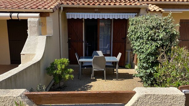 A house with a tiled roof, a covered patio with a table and chairs, and a small garden area.