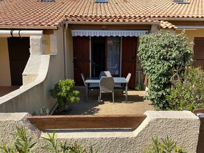 Ein Haus mit einem Ziegeldach, einer überdachten Terrasse mit Tisch und Stühlen sowie einem kleinen Garten.