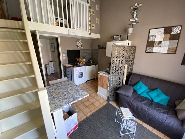 A compact living space with a kitchenette, a washing machine, a dining area, and a sofa with cushions.