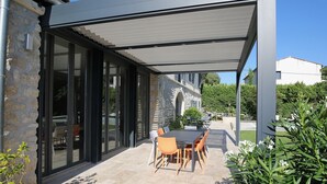 Eine überdachte Terrasse mit Tisch und Stühlen, Steinmauern und Blick auf ein Steinhaus im Hintergrund.