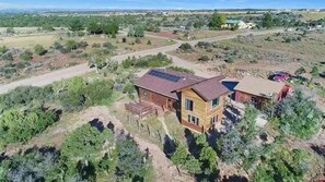 Una casa de dos pisos con techo solar, rodeada de un paisaje rural con poca vegetación y un camino de tierra.