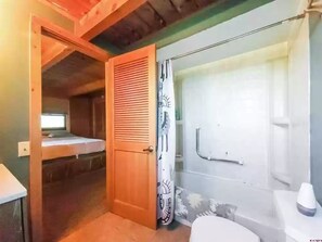 Una habitación pequeña y funcional con techo de madera, una cama individual, una puerta con panel decorativo, zona de ducha con mampara de vidrio y un lavabo blanco.