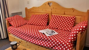 A wooden bed with a red and white heart-patterned bedspread, a small table with a mug, and a wall-mounted lamp. - Cozy 2-Room Apartment for 6 with Balcony, Near Slopes, Pet-Friendly (Risoul)