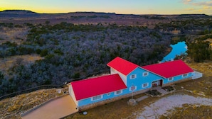 A two-story house with a red roof and blue walls, surrounded by a natural landscape with a river and hills. - Spacious Getaway ~ 12 Acres, Views, & Hot Tub! (Strawn)