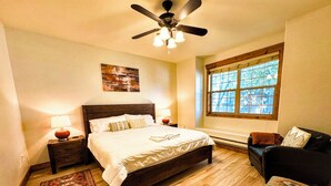 A bedroom with a wooden floor, a bed with white bedding, a ceiling fan, a window with blinds, and a nightstand with a lamp. - Mountain Chic Condo Near Grand Targhee and Driggs (Driggs)