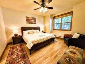 Quarto com piso de madeira, cama com roupa de cama branca, ventilador de teto, janela com persianas e mesa de cabeceira com luminária.