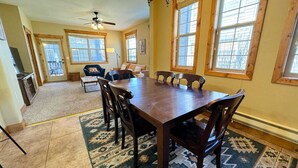 A spacious dining area with a large wooden table and chairs, a ceiling fan, and large windows with wooden frames.