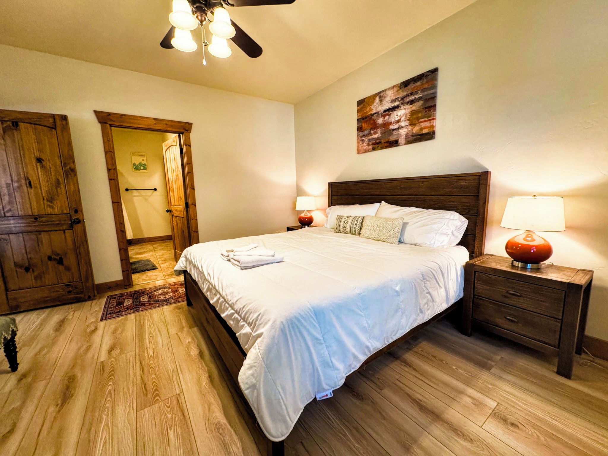 Ein Schlafzimmer mit Holzboden, einem Bett mit weißer Bettwäsche, einem Holz-Kopfteil, zwei Nachttischen mit Lampen und einem Deckenventilator.