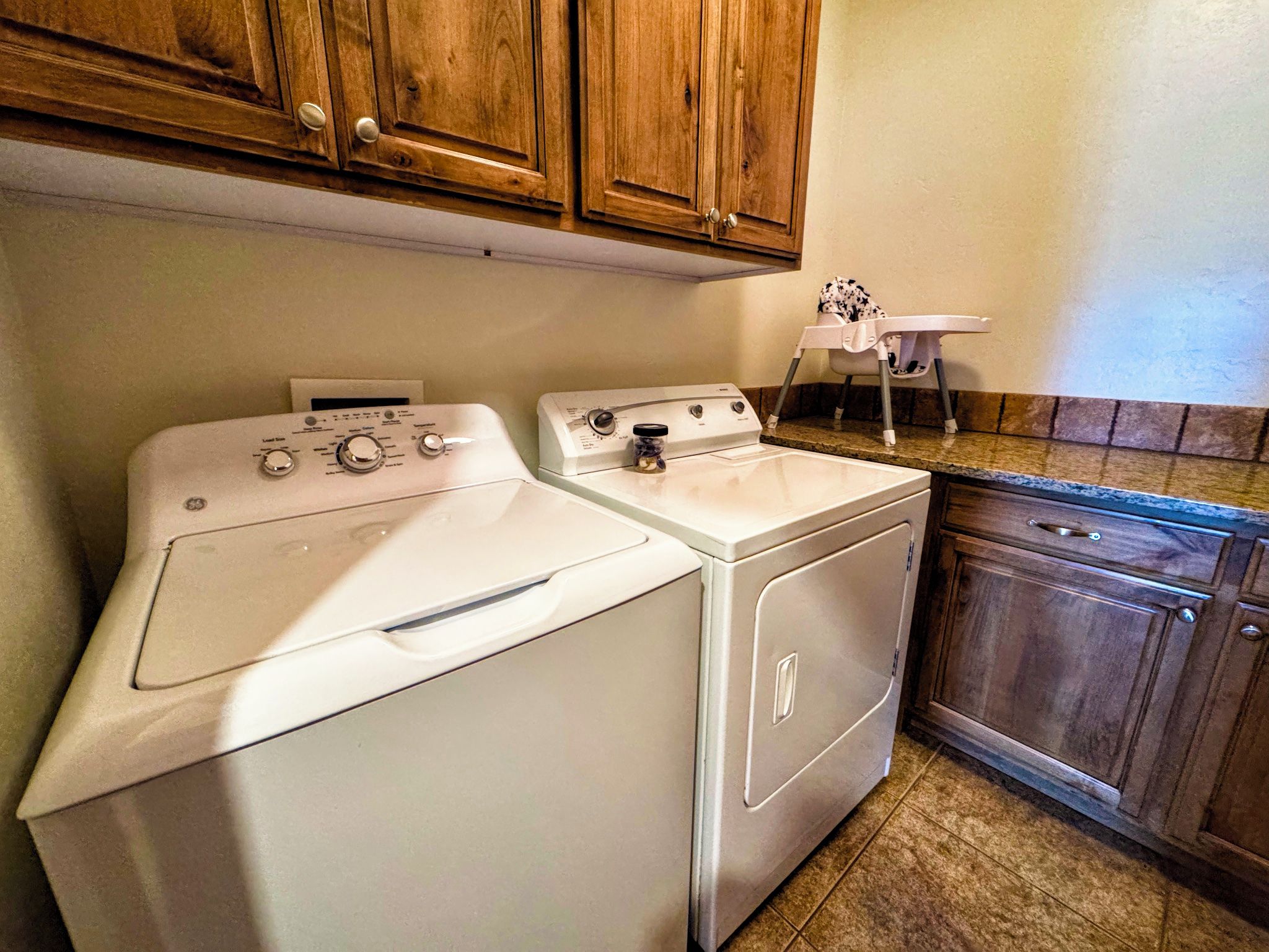 A laundry area with a front-loading washer and dryer, wooden cabinets above, and a small chair.