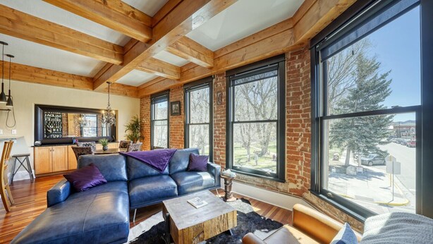 A modern living room with a leather sofa, wooden beams, large windows, and a kitchen area visible through the glass.
