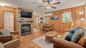 A cozy living room with a fireplace, wooden walls, a ceiling fan, and a dining area.