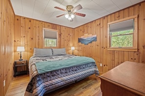 Un dormitorio con paneles de madera, una cama, dos mesitas de noche, un ventilador de techo y una ventana.